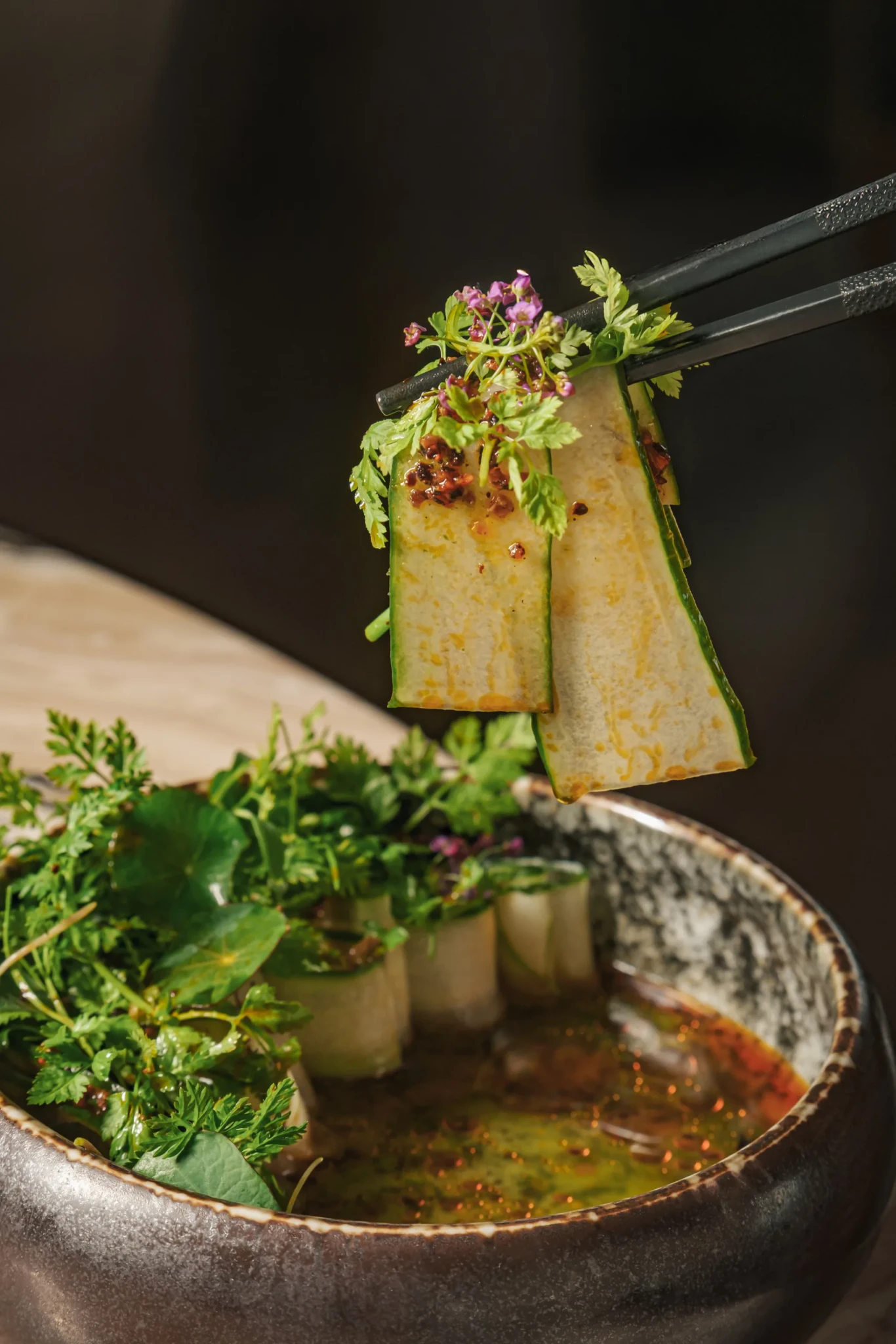 Chopsticks lift thin cucumber slices topped with purple flowers, herbs and chilli flakes over a bowl of dressing at CÉ LA VI Singapore