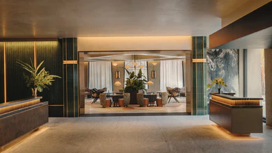 Hotel lobby with warm lighting, marble reception desks, potted plants and a lounge seating area beyond glass doors