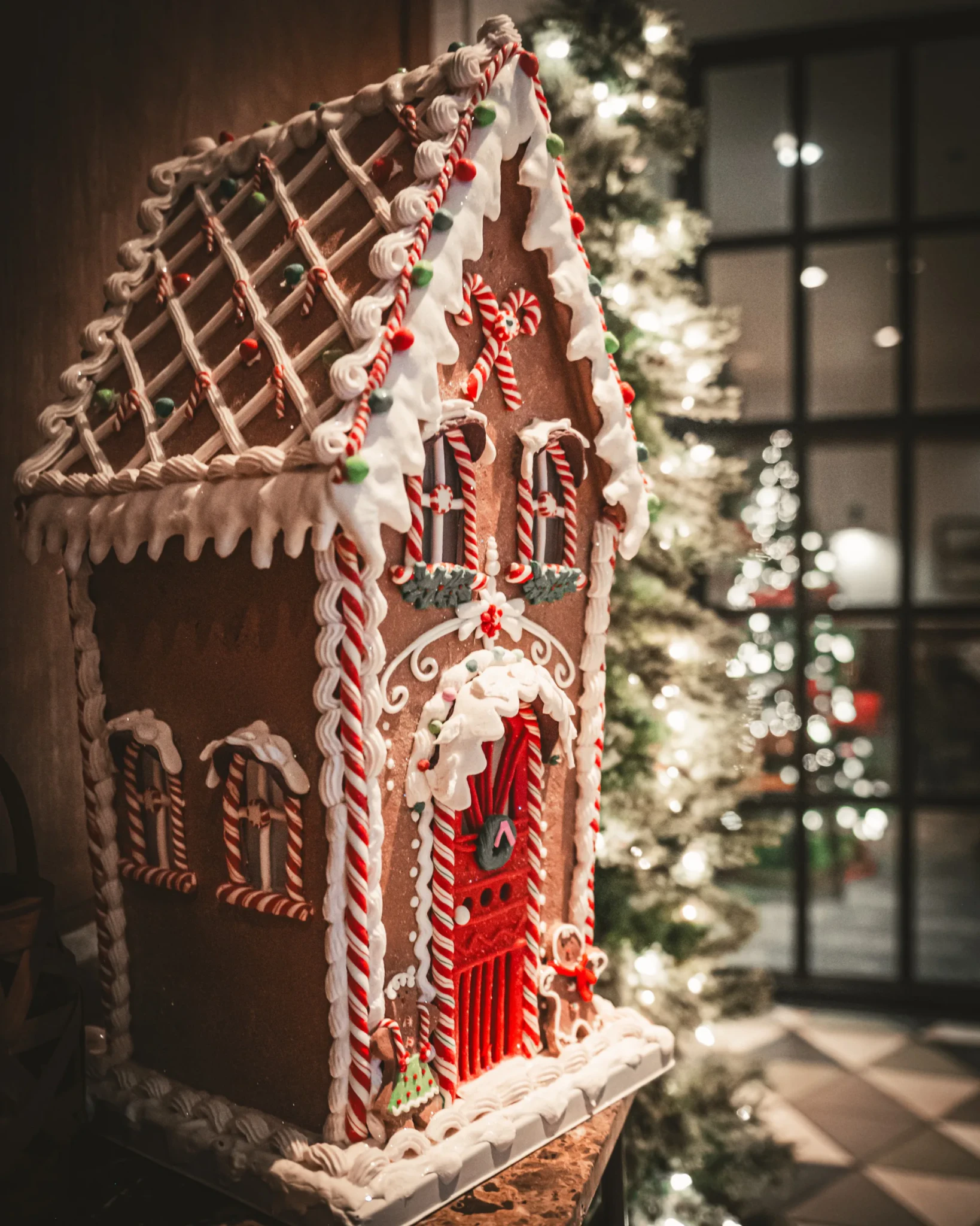 A gingerbread house decorated with white icing, candy canes and red and green gumdrops in front of a lit Christmas tree