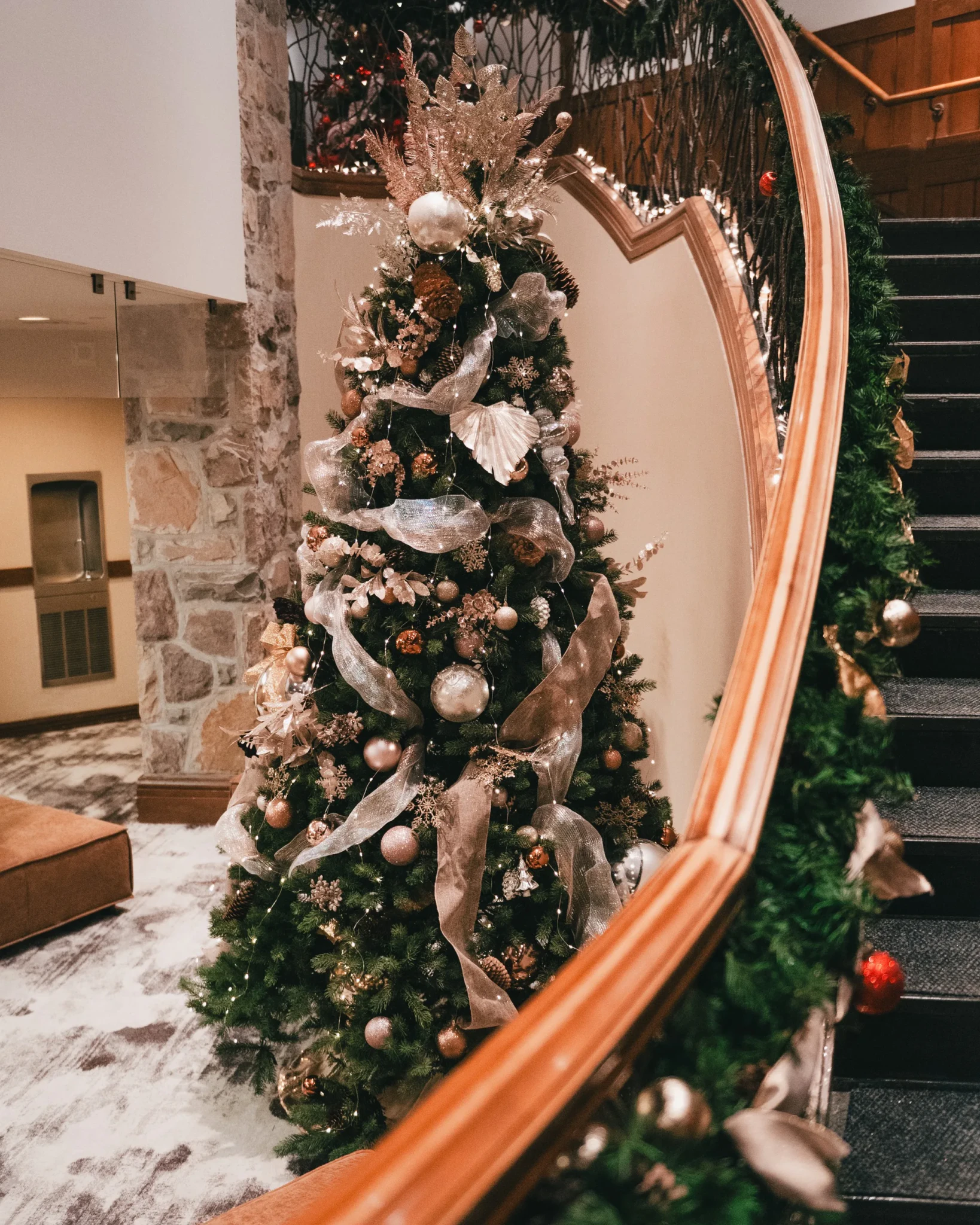 A Christmas tree in silver, champagne and rose-gold ornaments beside a curved wooden staircase in a Fairmont hotel lobby