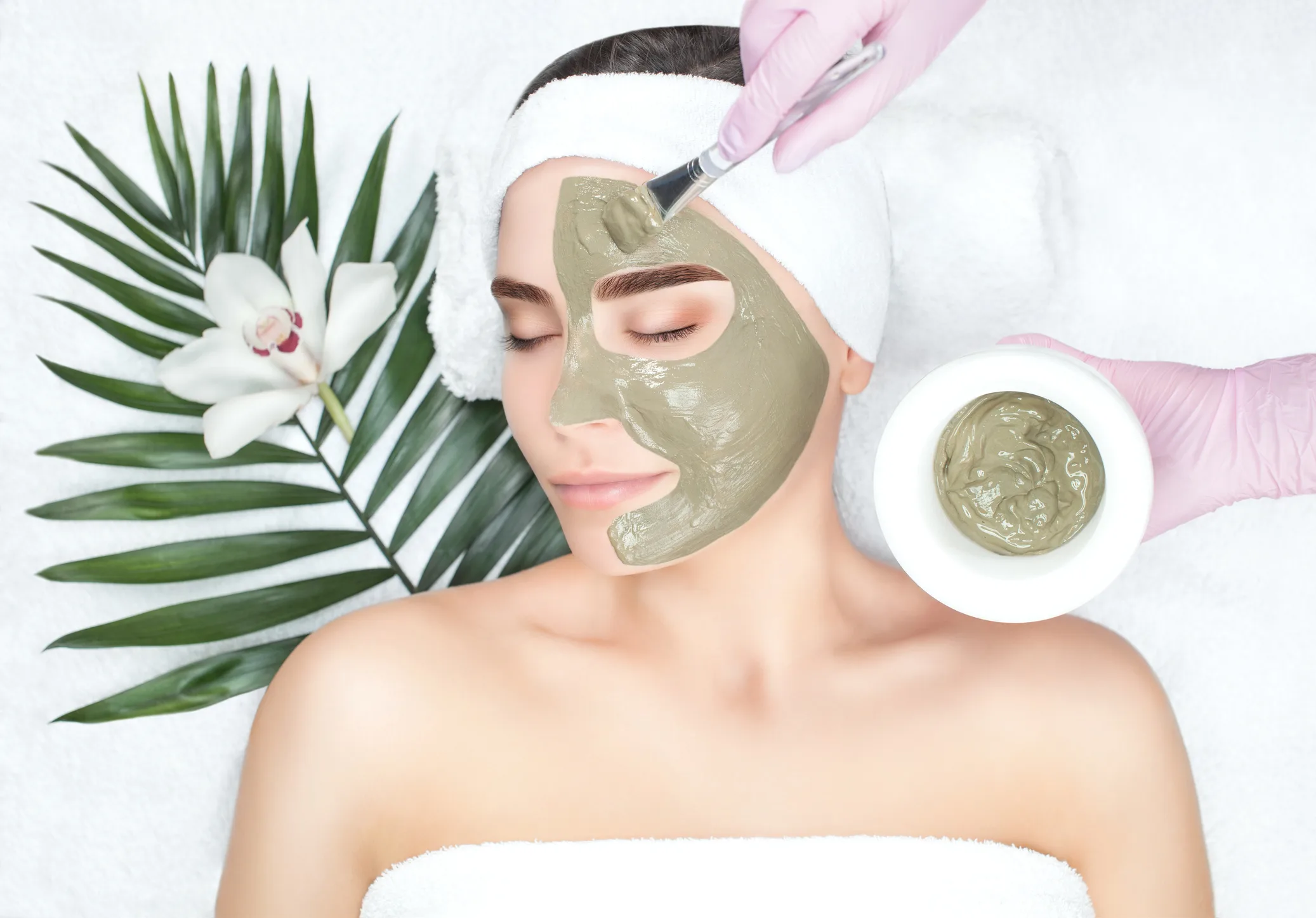 A woman receives a green clay facial mask applied with a brush, with a white orchid on a towel beside her