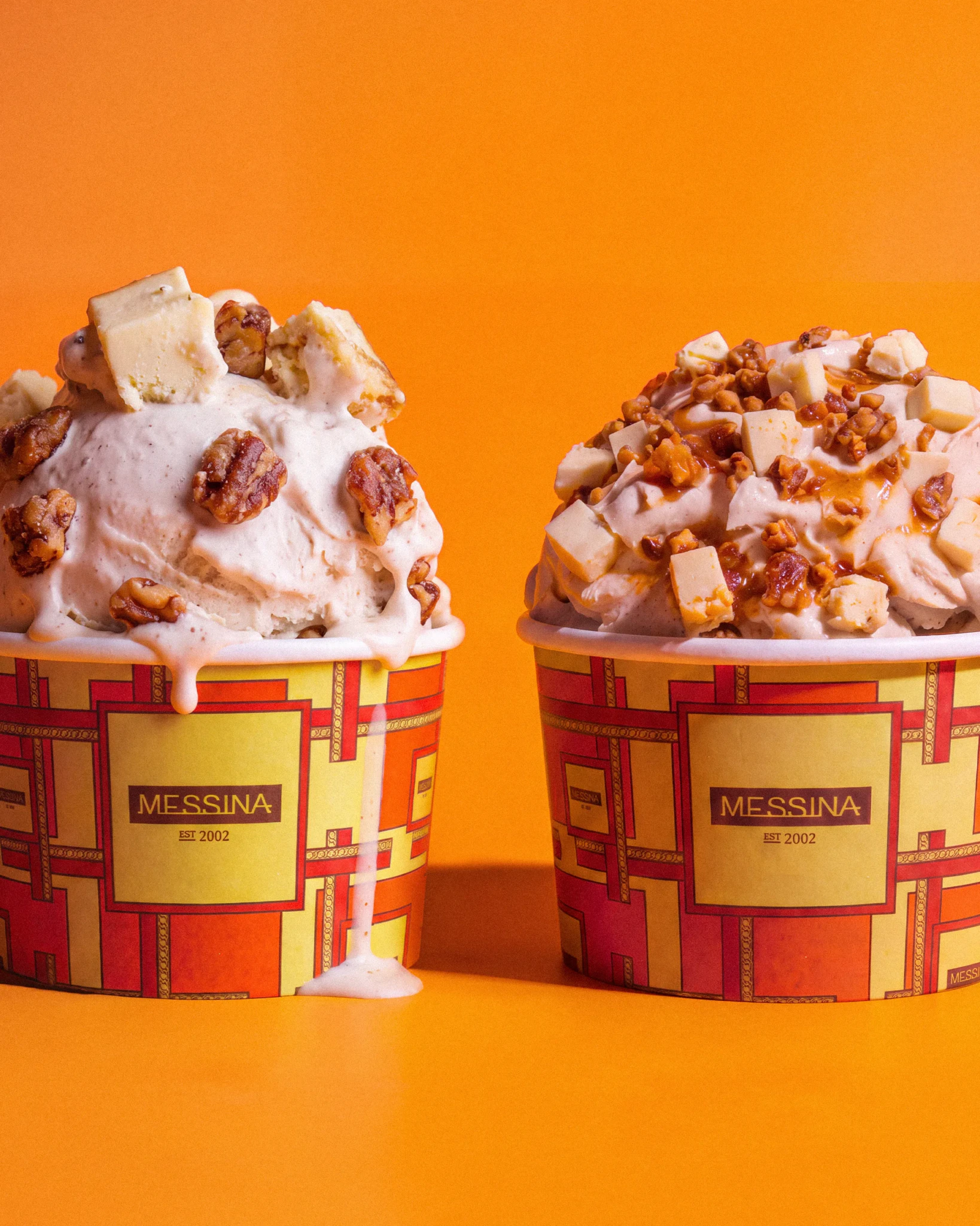 Two cups of Messina gelato topped with candied nuts and cheese chunks against a bright orange background
