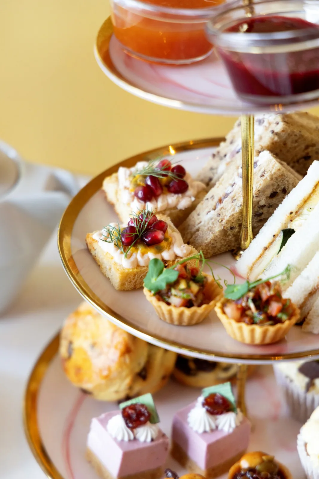 A tiered stand with smoked salmon mousse crostini topped with pomegranate seeds, savoury tartlets, sandwiches, raisin scones and pink petit desserts