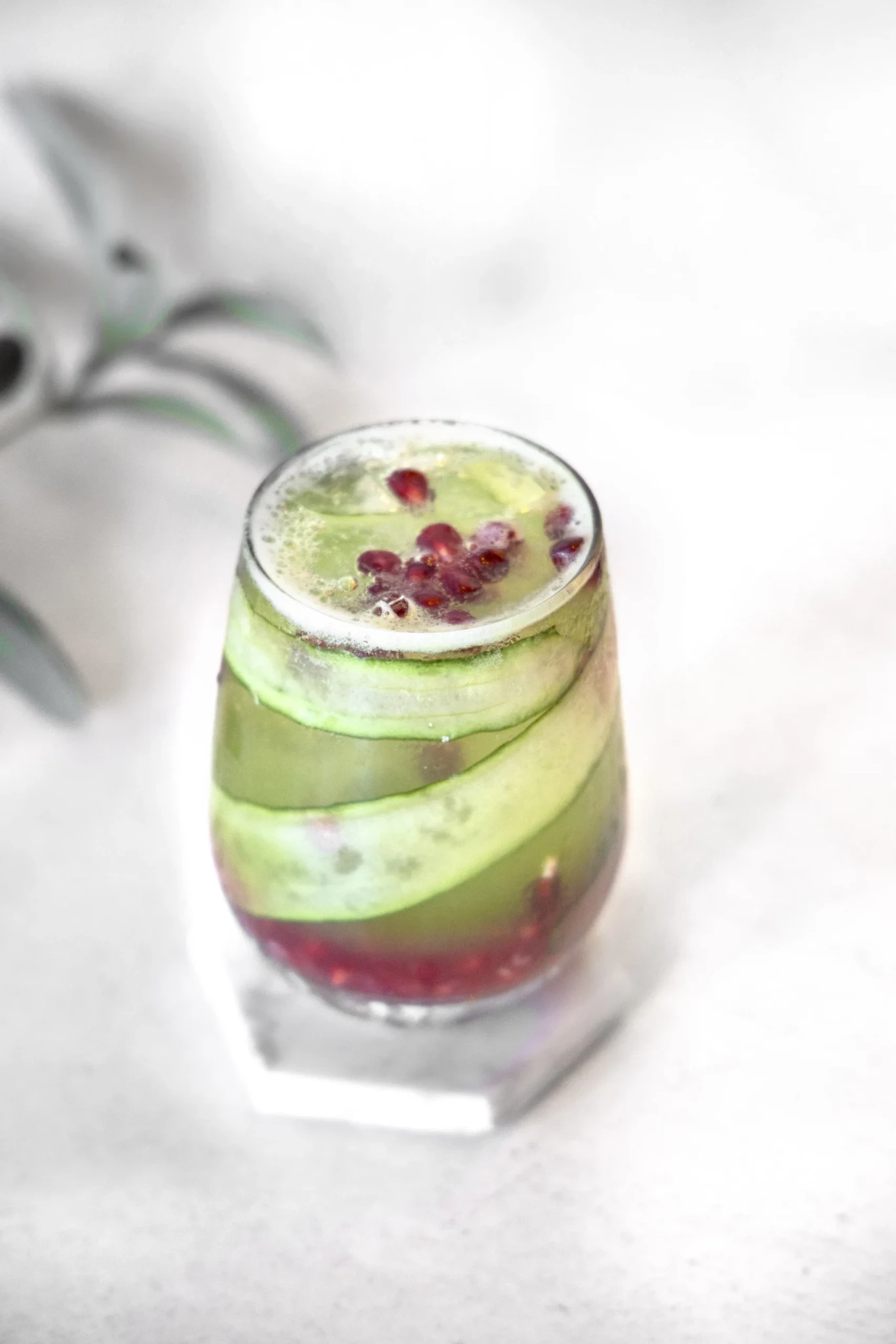 A glass of Holiday Fizz garnished with cucumber ribbons and pomegranate seeds on a white marble coaster
