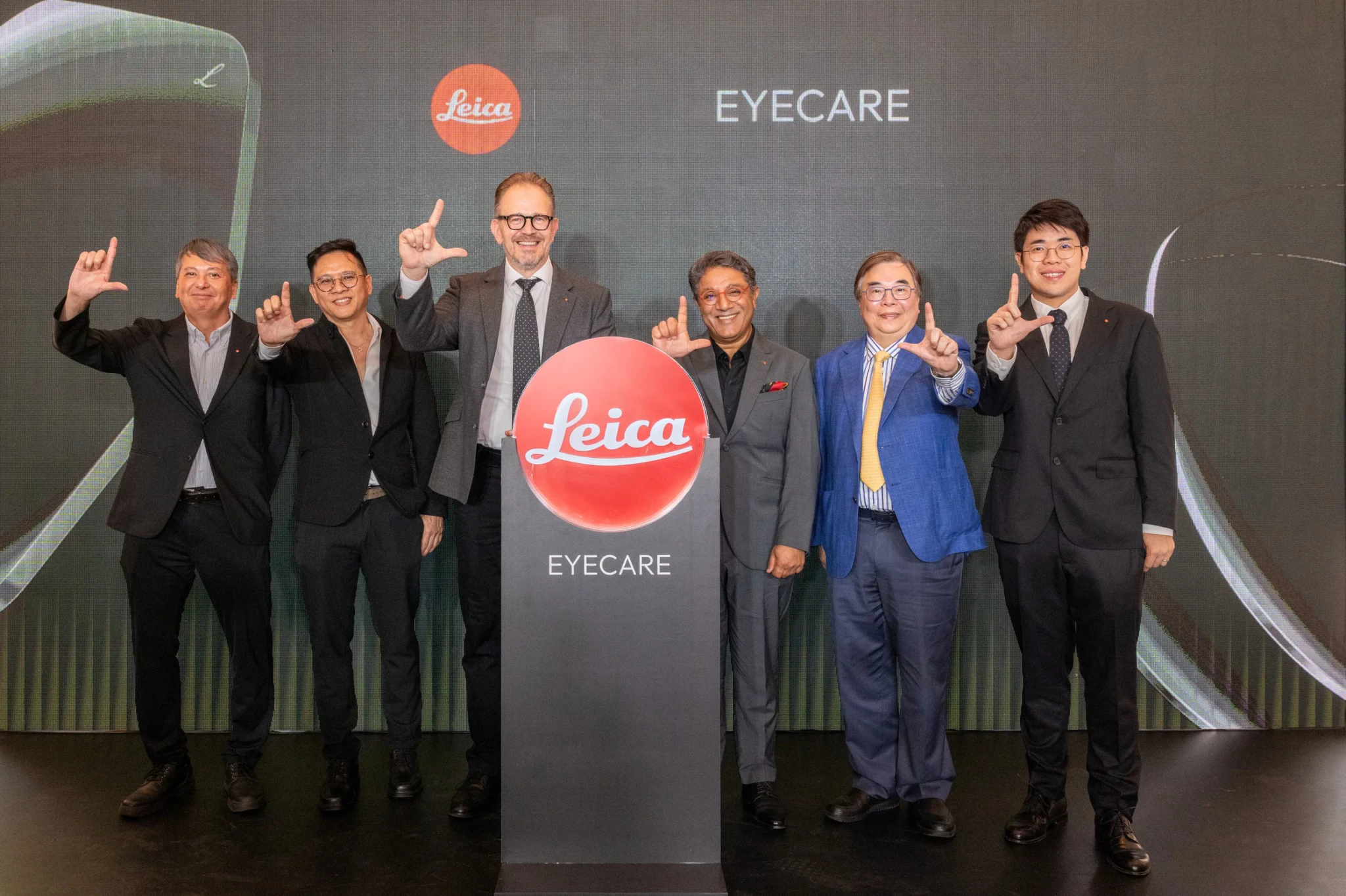 Six men pose behind a Leica EYECARE podium at the Singapore debut event, several making an L-shape gesture with their fingers in front of a screen reading EYECA