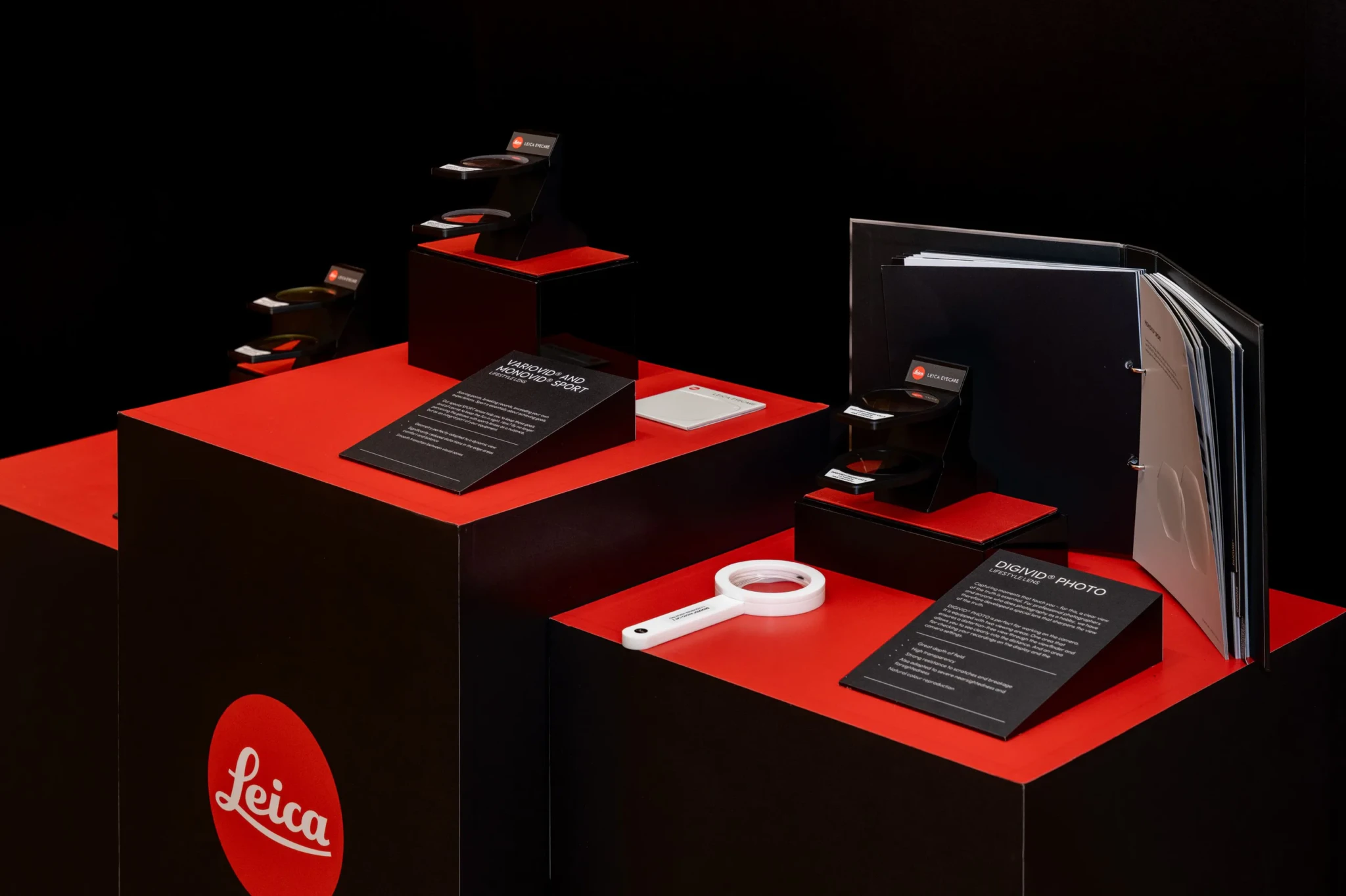 Leica Eyecare display on red and black plinths showing the VARIOVID and MONOVID sport lens samples and the DIGIVID PHOTO lens with its magnifier and sample book