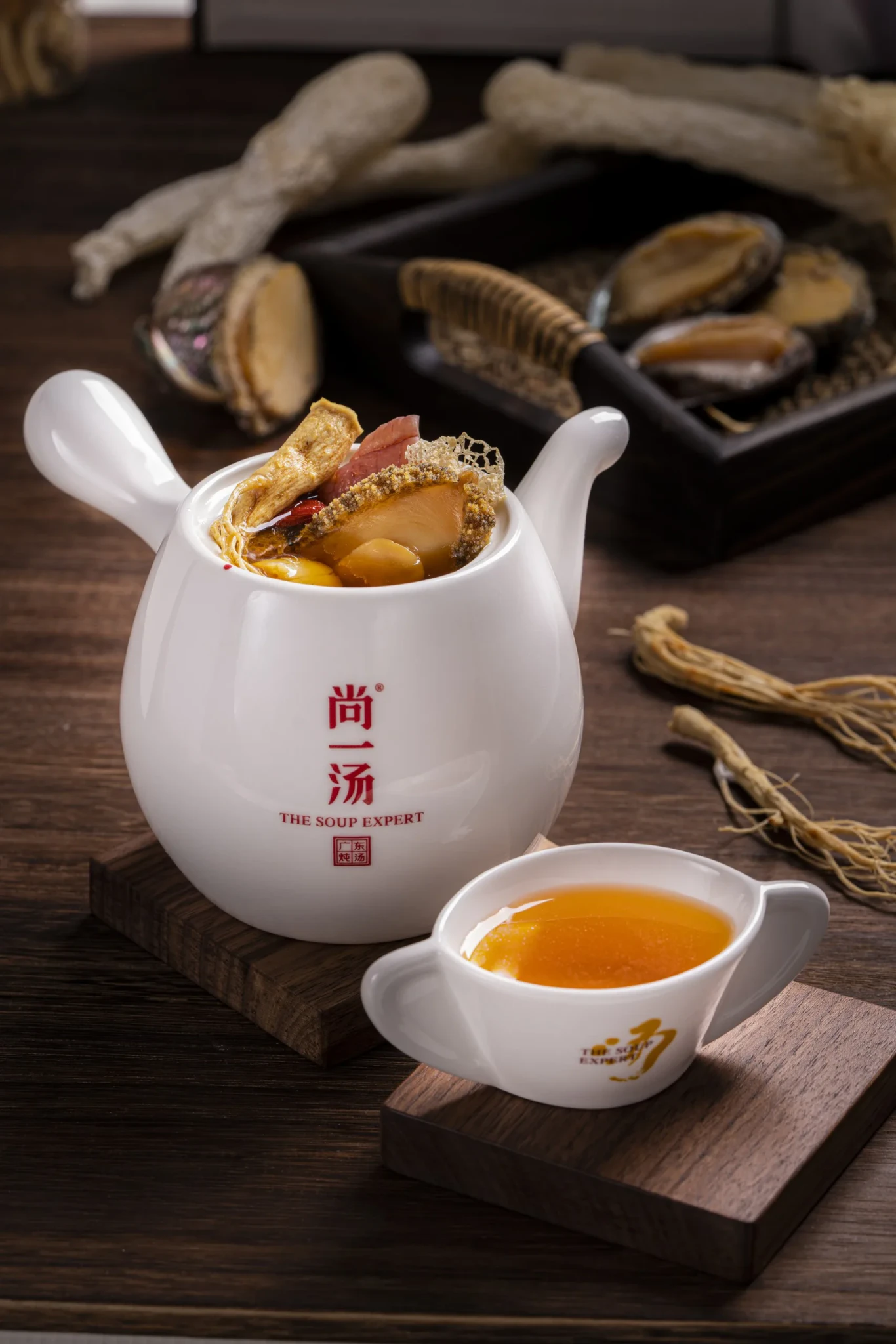 A white ceramic pot of Abalone Ginseng Soup with a small cup of amber broth, dried abalone and ginseng roots behind