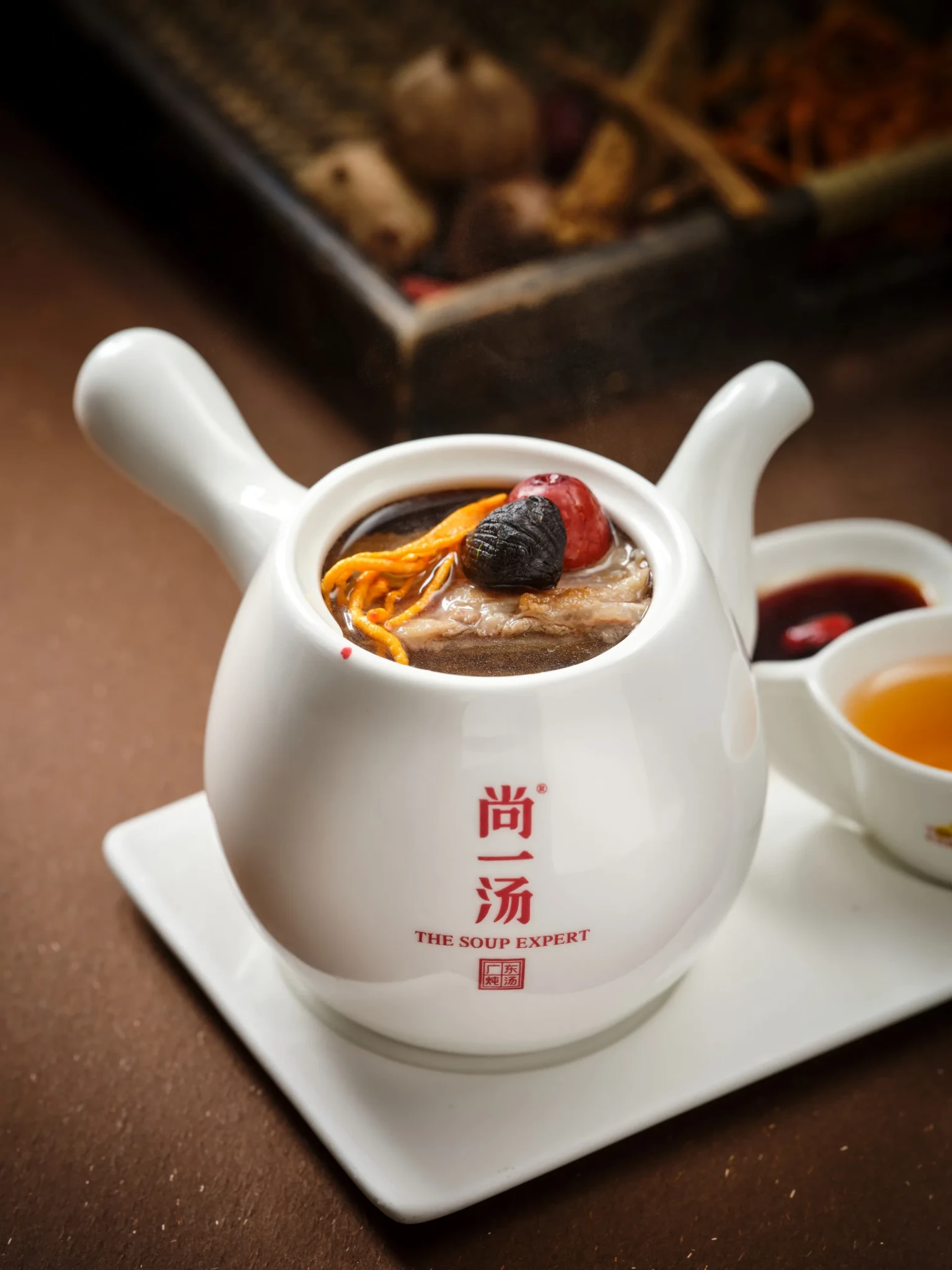 Black Garlic Beef Bone Soup in a white teapot-style pot topped with cordyceps, a black garlic clove and a red date