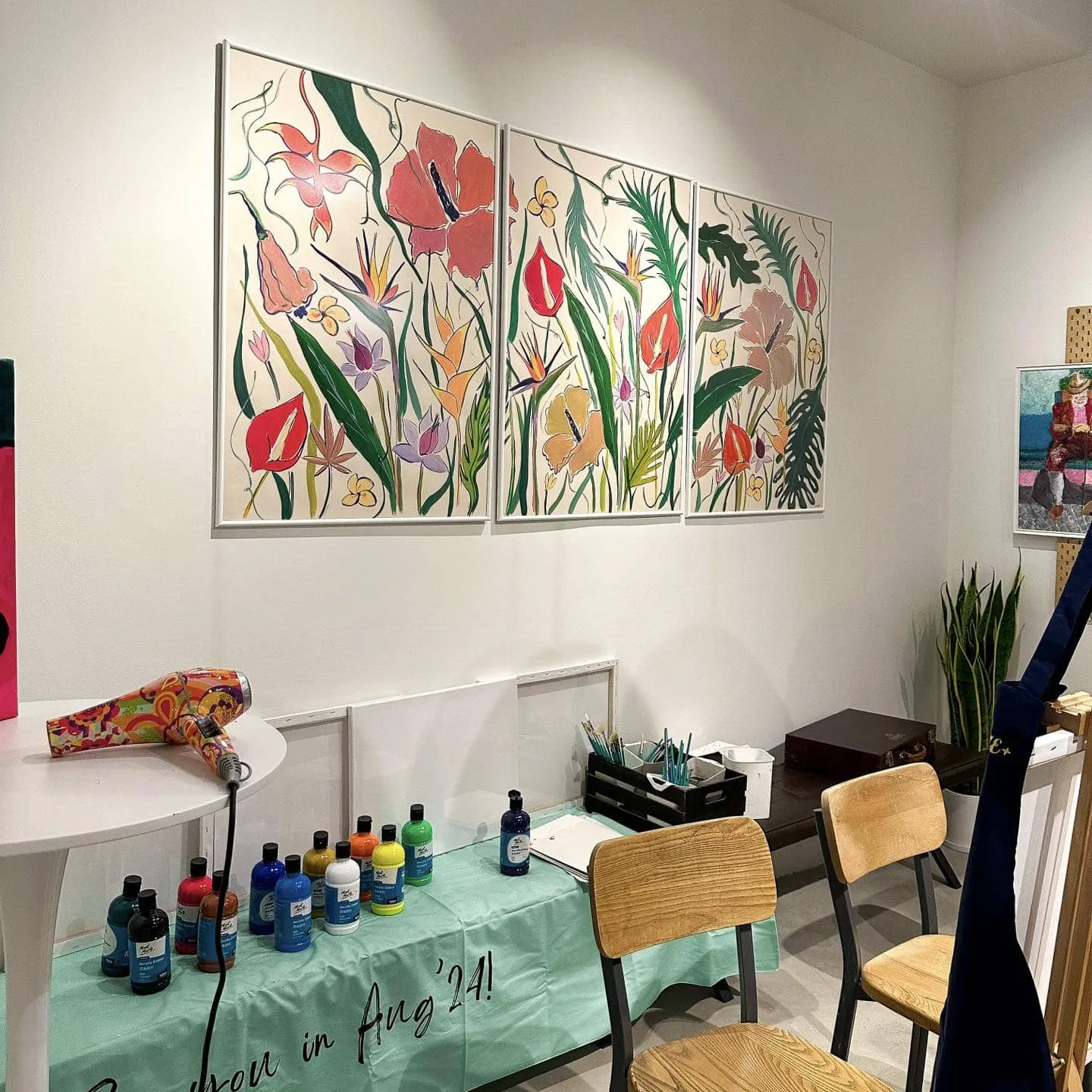 A painting table at Coze+ laid with acrylic paint bottles and a turquoise cloth, under three framed floral paintings